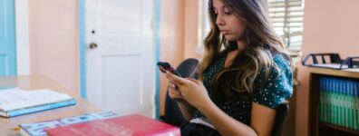 A teenage girl sits in a classroom, focused on her smartphone among books and educational materials.